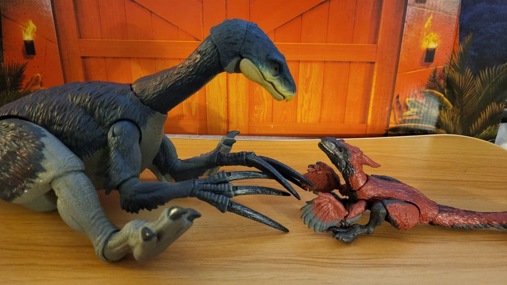 Therizinosaurus sitting down, facing towards the right, side view, alongside the Hammond Collection Pyroraptor, also sitting down, facing it to the right of it
