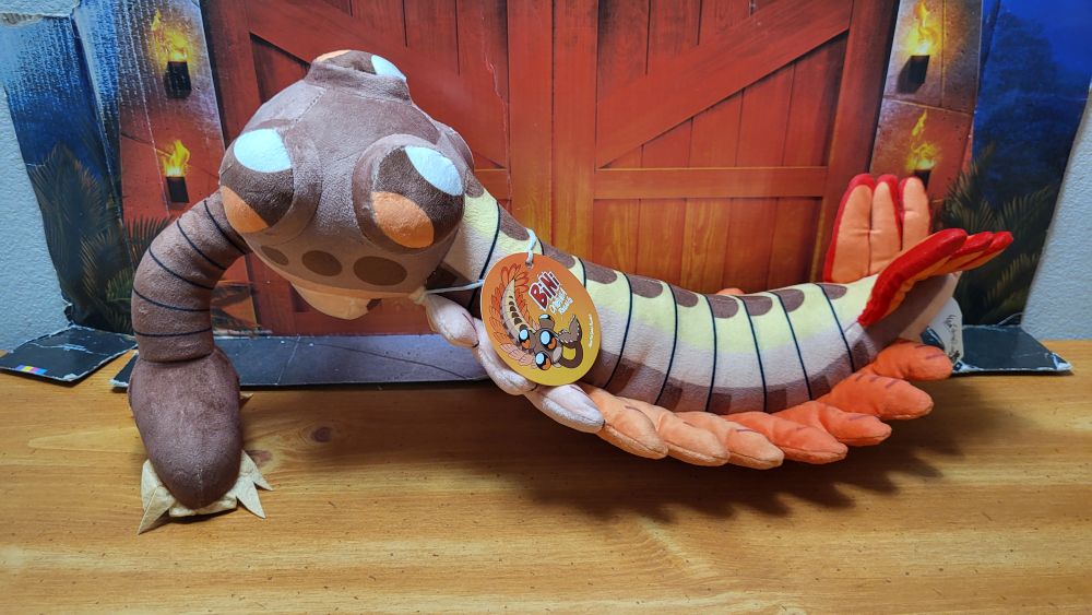 A side view of the plush, with the head facing the left side, and the tag is in view showing the front of it with the artwork by Saureal